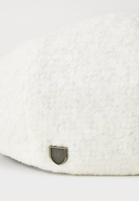 Beret de lana blanca con una superficie texturizada, que presenta un pequeño emblema metálico en forma de escudo con un diseño de rayas en el lateral.