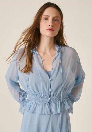 Femme aux longs cheveux portant un chemisier bleu clair transparent avec boutons, taille cintrée et ourlet à volants, porté sur un haut et une jupe assortis.