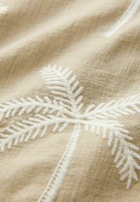 Tessuto di lino ricamato di colore beige con motivi bianchi a felce. La texture è morbida con un sottile motivo a griglia.