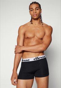 Black boxer shorts with a white elastic waistband featuring "ellesse" logo. Model stands with arms crossed, showcasing the shorts' fit.