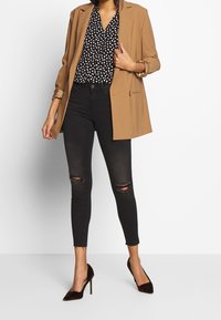 Blazer marrón sobre una blusa de lunares en blanco y negro, combinado con jeans ajustados negros con roturas en las rodillas y zapatos de tacón oscuros.