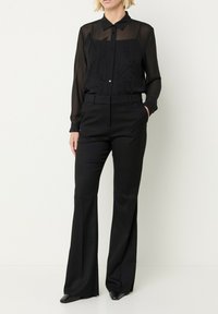 Donna che indossa una blusa nera trasparente a maniche lunghe con ricami floreali e pantaloni neri a vita alta e gamba larga, in piedi contro un muro bianco.