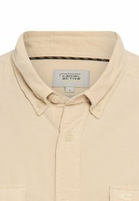 Beige ribfluwelen knoop-overhemd met een kraag, voorzien van een borstzak en een subtiele tekstuurpatroon. Merkenlabel zichtbaar binnen de kraag.
