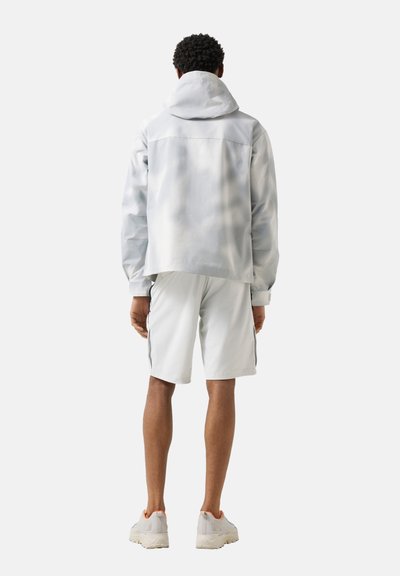 Mężczyzna ubrany w jasnoszarą kurtkę z kapturem, pasujące krótkie spodenki z bocznymi paskami oraz jasne sportowe buty, stojący tyłem na jednolitym tle.