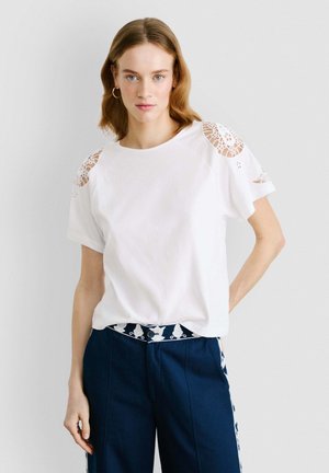 White short-sleeve t-shirt with lace detailing on shoulders, relaxed fit, and round neckline, paired with navy pants and patterned belt.