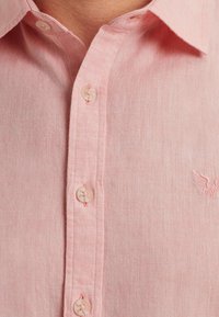 Camisa de lino rosa claro con cuello abotonado, con cuatro botones y un logo bordado en rojo en el lado izquierdo.