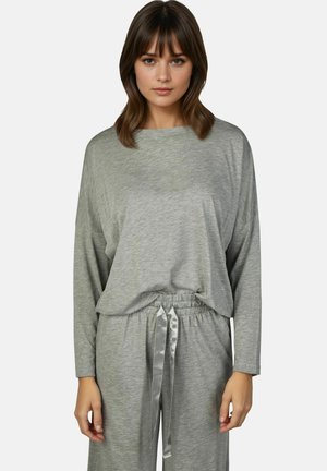 Vrouw met bruin haar die een losse grijze longsleeve en bijpassende grijze broek met trekkoord draagt tegen een effen achtergrond.