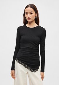 Schwarzes Langarmtop aus strukturiertem Stoff, mit Spitzenbesatz am Saum und figurbetonter Silhouette. Kombiniert mit hellen Hosen.