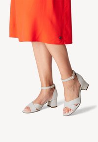Hvide læder sandaler med blokhæl og ankelrem, der har fletdetalje og åben tå-design, parret med en lys orange kjole.