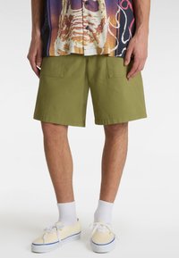 Olivengrønne shorts med løs pasform, fremstillet af bomuld. Har to store forlommer og en lige kant. Kombination med beige sneakers og hvide sokker.