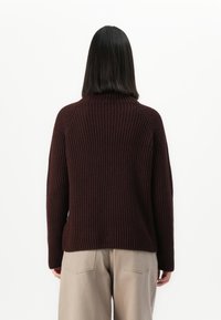 Eine Person mit glattem schwarzen Haar, die einen dunkelbraunen gerippten Pullover und beige Hosen trägt, steht vor einem schlichten weißen Hintergrund.