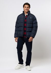 Marineblauwe pufferjack met horizontale naden, hoge kraag en een logo op de borst. Draag het over een gestreept rood en marineblauw shirt, gecombineerd met donkere jeans.