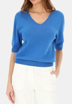 Kobieta w jasnoniebieskim swetrze z dzianiny z dekoltem w serek i rękawami do łokcia oraz białych spodniach z wysokim stanem, z ręką w kieszeni.