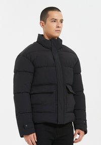 Veste en duvet noire avec un col montant, fermeture éclair à l'avant, grandes poches avant et texture matelassée. Tissu doux avec une finition mate.