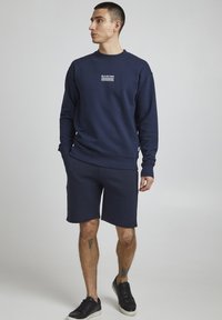 Hanorac bleumarin cu un model brodat alb pe piept, asociat cu şorturi asortate şi adidaşi negri. Material moale dintr-un amestec de bumbac.