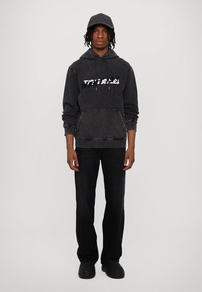 Black hoodie with white graphic text, front pocket, and drawstring hood. Dark jeans and black shoes. Fitted cap in matching color.