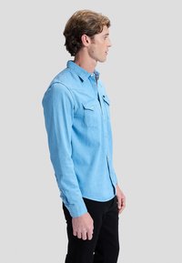 Jonge man in profiel met een lichtblauw denim button-up shirt en zwarte broek tegen een effen grijze achtergrond.