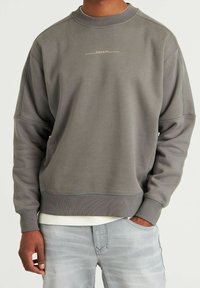 Sweatshirt gris à encolure ronde et épaules tombantes, arborant un logo discret en blanc sur la poitrine ainsi que des poignets et un ourlet côtelés.