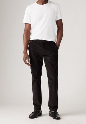 Man wearing a white short-sleeve t-shirt, black straight-leg pants, and black leather loafers, standing with one hand in pocket.