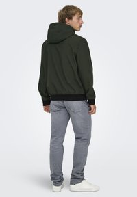 Giacca con cappuccio in tessuto verde scuro, caratterizzata da una vestibilità comoda, polsini a costine e orlo nero, abbinata a jeans grigio chiaro e scarpe da ginnastica bianche.