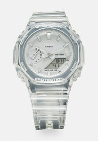 Montre Casio G-Shock avec un boîtier et un bracelet en résine transparentes. Présente un cadran blanc, trois sous-cadrans et un affichage numérique avec des étiquettes. Étanche jusqu'à 20 bars.