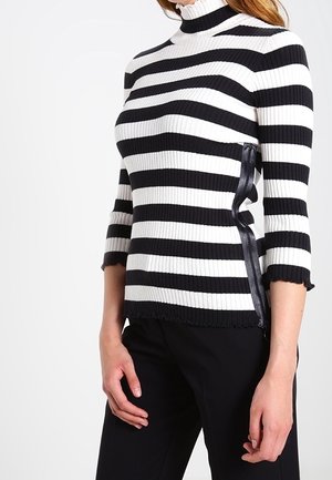 Femme portant un pull côtelé rayé horizontal noir et blanc avec manches trois-quarts et pantalon noir, debout devant un fond blanc.