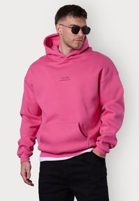 Haut à capuche rose avec une poche frontale, comportant un petit texte brodé. Confectionné en matériau doux avec une coupe décontractée et des poignets côtelés.