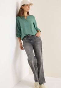 Hellgrün, langärmlige Bluse mit V-Ausschnitt und gerafften Manschetten, kombiniert mit grauen, geraden Jeans. Cremefarbene Sneakers vervollständigen das Outfit.