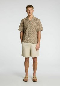 Camicia a maniche corte con bottoni, caratterizzata da un motivo geometrico marrone, abbinata a pantaloni corti beige e sandali beige, indossata da una persona in piedi.