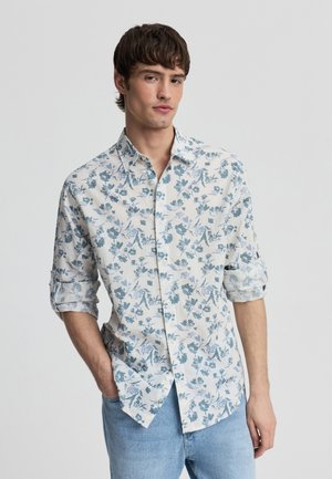 Młody mężczyzna w białej koszuli na guziki z niebieskim kwiatowym wzorem i podwiniętymi rękawami, w jasnoniebieskich dżinsach, z rękami w kieszeniach.