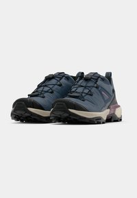 O pereche de pantofi de alergare trail albăstru și negri, cu tălpi robuste și sistem de șireturi rapide, înclinați ușor spre stânga pe un fundal simplu.