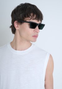 Gafas de sol negras con lentes oscuros; usadas por una persona con una blusa sin mangas blanca que presenta un sutil patrón texturizado.