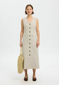 Beige linnedragt med V-hals, forreste knapper og to brystlommer. Sammen med en vævet taske og brune slip-on sko.