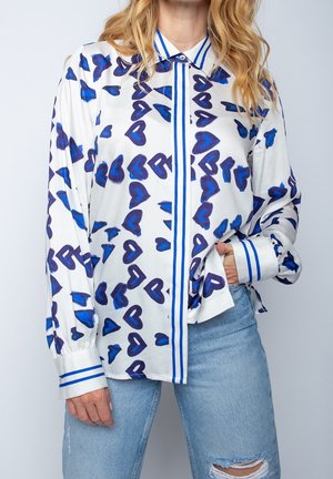 Wit hemd met knopen met blauwe en paarse hartpatronen, blauwe gestreepte accenten op de kraag en manchetten, gedragen met gescheurde lichtblauwe spijkerbroek.