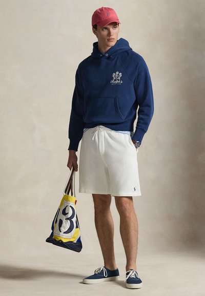 Μπλε σκούφο φούτερ με λογότυπο, λευκό σορτς, μπλε πάνινα παπούτσια και πολύχρωμη τσάντα ώμου με έντονο σχέδιο. Ανοιχτόχρωμο φόντο.