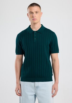 Dunkelgrünes Poloshirt mit kurzen Ärmeln, ribbeliger Textur, Drei-Knopf-Leiste und geradem Saum. Kombiniert mit hellen Jeans.