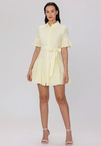 Robe à manches courtes et à boutons, de couleur jaune clair, avec un ourlet à volants en plusieurs niveaux et une ceinture en tissu. Froufrous verticaux et détails de col visibles.