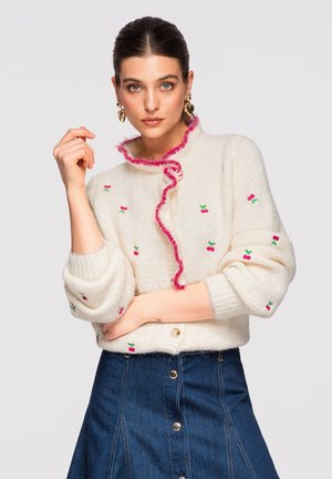 Kvinde iført en hvid, fluffy sweater med små rosa kirsebær og rosa flæsekanter, kombineret med en denimnederdel med knapper foran og guldbøjler.