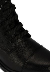 Nahaufnahme eines schwarzen strukturierten Lederstiefels mit dicken schwarzen Schnürsenkeln, die durch metallene Ösen gefädelt sind.