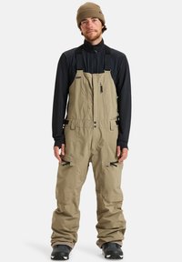 Bežna vodoodporna kombinezona z nastavljivimi naramnicami, žepom na zadrgo na prsih in stranskimi ventilacijskimi odprtinami, nosena preko črne majice z dolgimi rokavi in črnih čevljev.