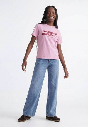 Chica sonriente con trenzas largas lleva una camiseta rosa de Montreal Quebec, vaqueros azules de pierna ancha y zapatos marrones, de pie contra un fondo blanco.