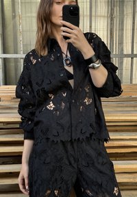Camicia di pizzo nero e pantaloni abbinati con intricati motivi floreali e ritagli. Gli orli sono sagomati, con chiusura frontale a bottoni e maniche morbide.