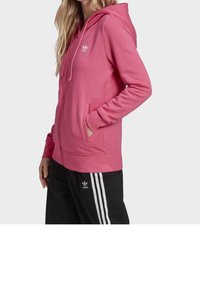 Růžová mikina na zip z měkké látky, s kapucí, předními kapsami a bílým logem Adidas na levé straně hrudi. Černé kalhoty s třemi pruhy.