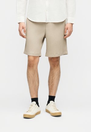 Bărbat purtând pantaloni scurți bej cu dungi, tricou alb, șosete negre și adidași albi cu talpă de gumă, stând în picioare pe un fundal simplu.