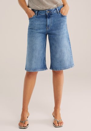 Naine kannab hele siniseid keskmise pikkusega teksapükse taskutega ees, millega on kombineeritud avatud varbaga kontsaga sandaalid ja kootud särk.