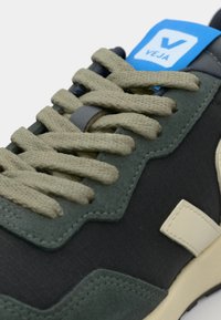 Sko med teksturert svart syntetisk overdel, mørkegrønn suede-detalj, beige logo og beige snøringer. Har en blå merkevaretag.