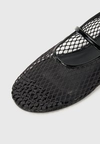Chaussure en mesh noir avec un bout rond et une bande noire brillante sur le cou-de-pied, dotée d'un design tissé ouvert et d'une semelle lisse.