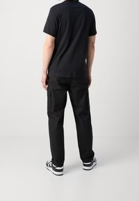 Man standing facing away wearing a black t-shirt, black cargo pants, and black and white sneakers on a plain white background.