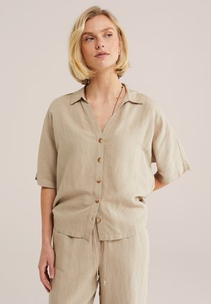 Femme aux cheveux blonds courts portant une chemise beige à boutons et un pantalon assorti à cordon, sur un fond uni.