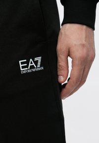 Tuvplāns ar roku pie melnas drēbes, uz auduma ir iespiests balts "EA7 Emporio Armani" logotips.